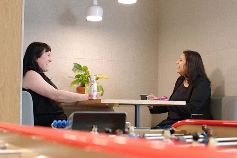 Two women engaged in conversation while sitting at a table in a bright office setting, surrounded by paperwork and laptops.