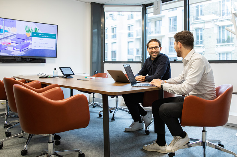 Two men discussing ideas at a table in an office, with laptops in the center of their meeting.