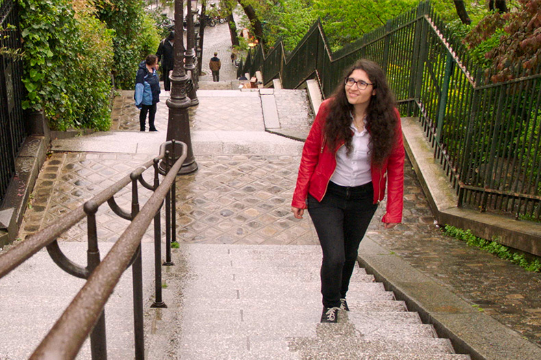 A person with long hair and glasses, wearing a red jacket and black pants, walks up a stone staircase outdoors, surrounded by greenery and other people in the background.