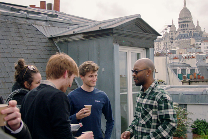 A group of people are gathered on a rooftop with drinks, engaged in conversation. A historic domed building is visible in the background.